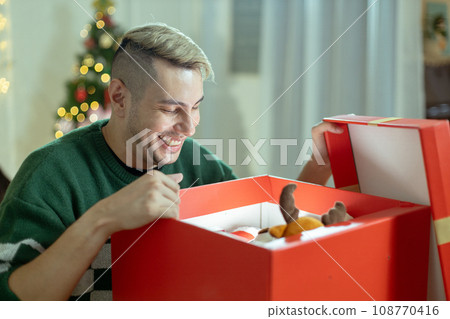 Christmas day concept. handsome man sitting surprised and opening gift box with christmas tree  Christmas day concept. handsome man sitting surprised and opening gift box with christmas tree  108770416