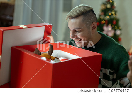 Christmas day concept. handsome man sitting surprised and opening gift box with christmas tree  Christmas day concept. handsome man sitting surprised and opening gift box with christmas tree  108770417