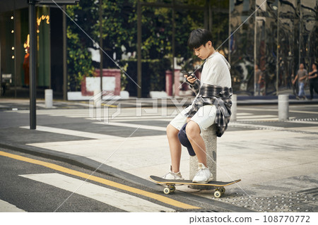 teenage asian skateboarder kid looking at mobile phone while resting teenage asian skateboarder kid looking at mobile phone while resting 108770772