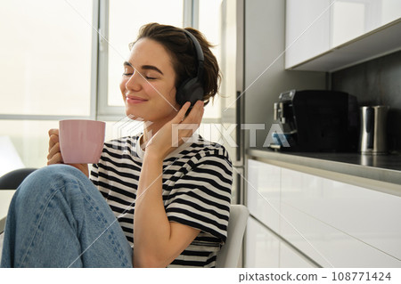 Lifestyle and music concept. Happy smiling woman with cup of tea, sits in kitchen and touches headphones on her head, listens to music with pleased relaxed face Lifestyle and music concept. Happy smiling woman with cup of tea, sits in kitchen and touches headphones on her head, listens to music with pleased relaxed face 108771424