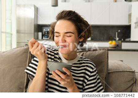 Portrait of happy brunette woman sitting on sofa and enjoying her breakfast, eating cereals, healthy granola in bowl, spending time in living room 108771442