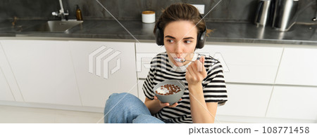 Portrait of carefree brunette woman, eating her cereals, listening to music, having breakfast in the kitchen 108771458