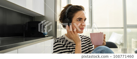 Close up portrait of smiling brunette woman, student drinks her tea and listens music or ebook in headphones, sits in kitchen and relaxes Close up portrait of smiling brunette woman, student drinks her tea and listens music or ebook in headphones, sits in kitchen and relaxes 108771460