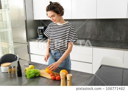 Portrait of modern young woman standing in the kitchen with vegetables, chopping them on counter, holding knife, cooking meal, vegetarian food, concept of healthy lifestyle an diet 108771482