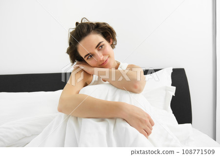 Portrait of smiling, happy young woman with sensual face, relaxing in bed, waking up, sitting with dreamy expression, resting at home 108771509