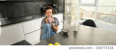 Cute brunette woman, student eating breakfast in morning, drinking hot tea, smiling and looking pleased 108771528