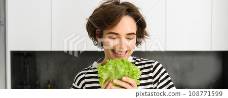 Portrait of happy and healthy young woman, following her diet, posing with lettuce leaf and smiling, cooking in the kitchen, vegetarian loves her vegetables Portrait of happy and healthy young woman, following her diet, posing with lettuce leaf and smiling, cooking in the kitchen, vegetarian loves her vegetables 108771599