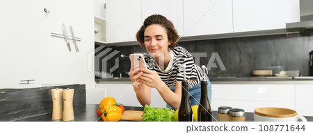 Woman bending on kitchen counter with smartphone, looking for healthy recipe online on mobile phone, standing next to chopping board and vegetables, cooking ingredients 108771644
