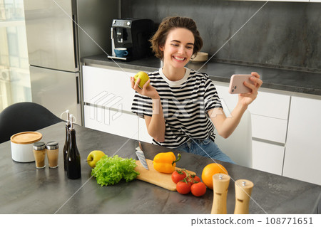 Portrait of beautiful young woman, food blogger recording video of her cooking in the kitchen, showing meal ingredients, preparing a meal on camera, using smartphone for live streaming Portrait of beautiful young woman, food blogger recording video of her cooking in the kitchen, showing meal ingredients, preparing a meal on camera, using smartphone for live streaming 108771651