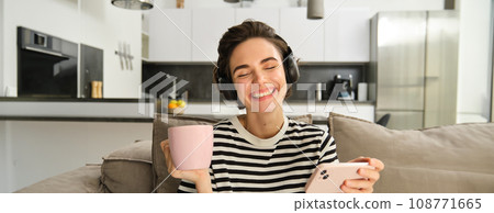 Carefree female model in headphones, sitting on sofa with smartphone and cup of tea, watching videos in wireless earphones on mobile phone app, resting at home 108771665