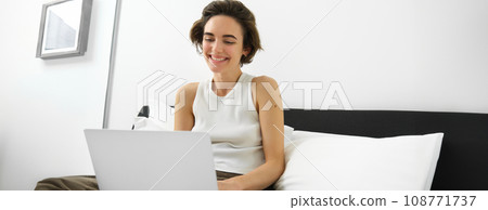 Image of smiling, happy young woman in bedroom, sitting on bed with laptop, laughing and looking at computer screen, working from home, freelancing 108771737
