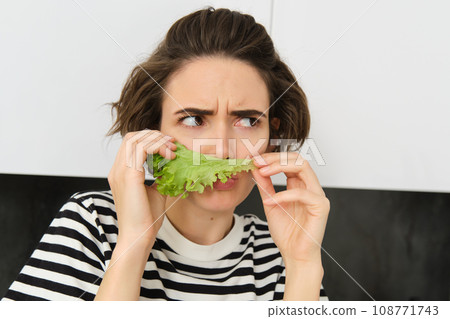 Portrait of woman frowning and grimacing, smelling fresh lettuce leaf with displeased face, standing in the kitchen, dislike vegetables Portrait of woman frowning and grimacing, smelling fresh lettuce leaf with displeased face, standing in the kitchen, dislike vegetables 108771743