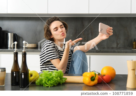 Cute young woman, lifestyle blogger cooking, sitting in the kitchen with vegetables and chopping board, taking selfie on her new smartphone, sending kisses at mobile camera 108771859
