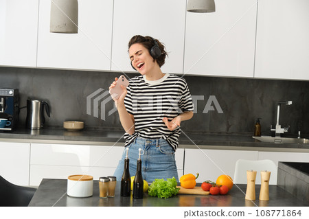 Carefree woman cooking in the kitchen, singing along favourite song, listening music in wireless headphones, holding mobile phone, chopping vegetables, preparing healthy meal 108771864