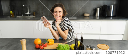 Happy smiling woman, sitting in the kitchen with smartphone, cooking salad, has chopping board, olive oil and vegetables on table Happy smiling woman, sitting in the kitchen with smartphone, cooking salad, has chopping board, olive oil and vegetables on table 108771912