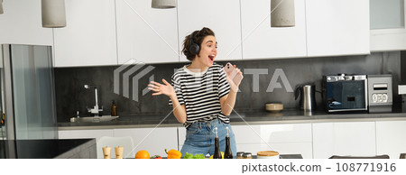 Portrait of woman singing and dancing while cooking food in the kitchen, holding smartphone, chopping vegetables on kitchen counter Portrait of woman singing and dancing while cooking food in the kitchen, holding smartphone, chopping vegetables on kitchen counter 108771916