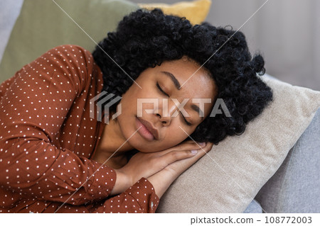 Young beautiful woman resting at home after work, African American woman sleeping napping on sofa in living room during daytime, day nap. 108772003