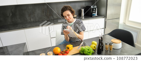 Portrait of woman writing down her recipe, making notes in notebook during cooking, sitting in the kitchen with chopping board and vegetables, preparing a meal Portrait of woman writing down her recipe, making notes in notebook during cooking, sitting in the kitchen with chopping board and vegetables, preparing a meal 108772023