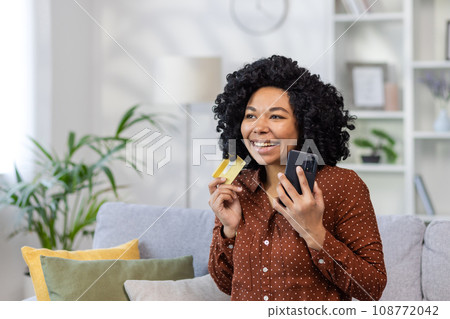 Joyful happy woman buyer online internet store, satisfied smiling sitting on the sofa in living room on house, holding a bank credit card in hands, using application on her phone, placing an order. Joyful happy woman buyer online internet store, satisfied smiling sitting on the sofa in living room on house, holding a bank credit card in hands, using application on her phone, placing an order. 108772042