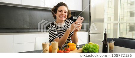 Portrait of brunette girl cooking food in the kitchen, searching recipes on social media app, holding mobile phone, standing near chopping board and vegetables, preparing healthy vegetarian meal Portrait of brunette girl cooking food in the kitchen, searching recipes on social media app, holding mobile phone, standing near chopping board and vegetables, preparing healthy vegetarian meal 108772126