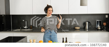 Portrait of happy girl cooking salad and listening to music in headphones, dancing while making a meal in the kitchen 108772140