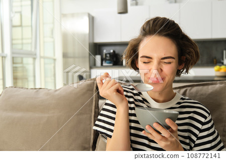 Portrait of young female model, student eating quick breakfast, holding bowl of granola or cereals with milk, sitting on sofa in living room Portrait of young female model, student eating quick breakfast, holding bowl of granola or cereals with milk, sitting on sofa in living room 108772141