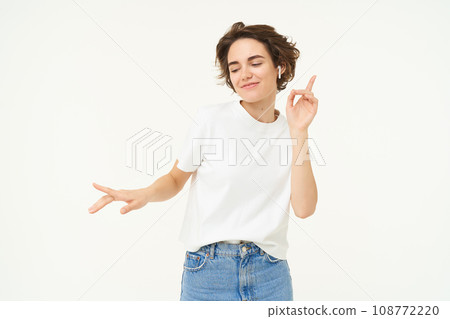 Portrait of carefree, happy young woman in wireless headphones, listens to music, dances to favourite song in earphones, posing over white studio background 108772220
