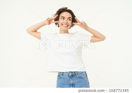 Happy smiling woman, showing peace, v-sign gesture and listening to music in wireless headphones, posing over white background 108772395