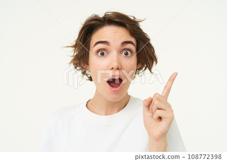 Excited girl has an idea, thinks of solution, pointing one finger and smiling, standing over white background 108772398