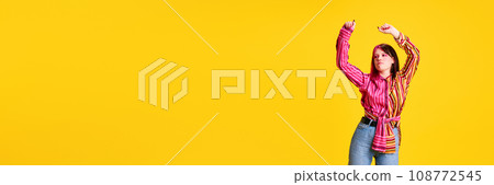 Young girl with pink hair, bright makeup and piercing, in colorful t-shirt dancing against vivid yellow studio background Young girl with pink hair, bright makeup and piercing, in colorful t-shirt dancing against vivid yellow studio background 108772545