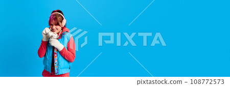 Young girl with bright makeup, in warm winter clothes eating ice cream against blue studio background. Winter vacation Young girl with bright makeup, in warm winter clothes eating ice cream against blue studio background. Winter vacation 108772573