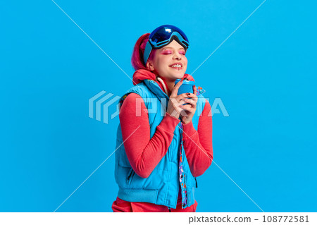 Warming up with hot tea. Smiling young girl with bright makeup wearing winter warm clothes against blue studio background Warming up with hot tea. Smiling young girl with bright makeup wearing winter warm clothes against blue studio background 108772581