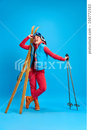 Young girl in colorful winter clothes standing with skis against blue studio background. Active holidays Young girl in colorful winter clothes standing with skis against blue studio background. Active holidays 108772583