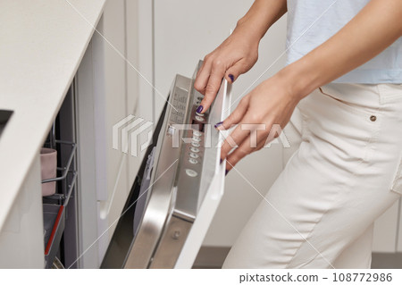female hand turning on dishwasher machine in kitchen, woman choosing mode program on control panel of dishwasher. female hand turning on dishwasher machine in kitchen, woman choosing mode program on control panel of dishwasher. 108772986