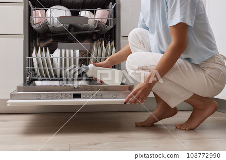 female hand pours rinse aid into the dishwasher compartment in modern white kitchen female hand pours rinse aid into the dishwasher compartment in modern white kitchen 108772990