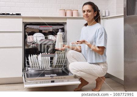 woman pours rinse aid into the dishwasher compartment in modern white kitchen woman pours rinse aid into the dishwasher compartment in modern white kitchen 108772991