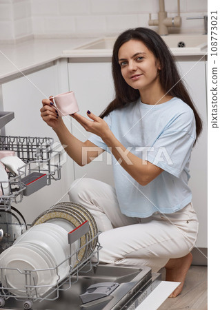 pretty woman unloading from open automatic built-in dishwasher machine with clean utensils inside in modern white kitchen. 108773021