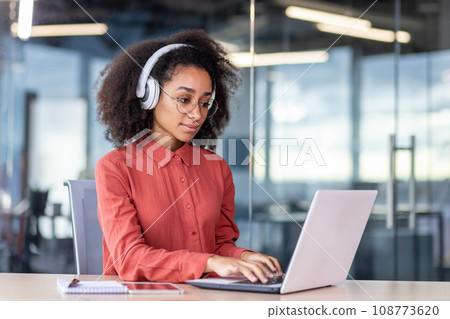 Serious concentrated woman working with laptop inside office, businesswoman thinking with headphones typing on keyboard, programmer developer listening to audio podcast, training course. Serious concentrated woman working with laptop inside office, businesswoman thinking with headphones typing on keyboard, programmer developer listening to audio podcast, training course. 108773620