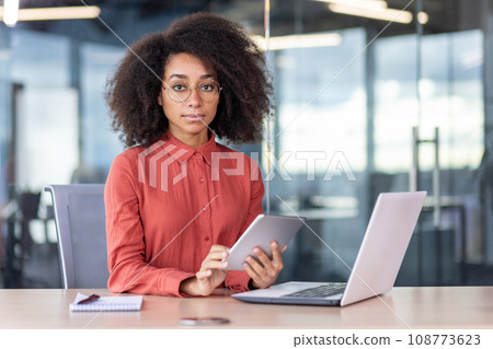 Portrait of confident focused woman at workplace, business woman looking thoughtfully at camera, holding tablet computer, sitting at desk with laptop, programmer developer, testing new programs. 108773623