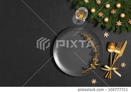 Christmas table setting with fir branches, golden decorations on black background. Empty black plate for design. Happy new year. Space for text. Top view, flat lay. Christmas table setting with fir branches, golden decorations on black background. Empty black plate for design. Happy new year. Space for text. Top view, flat lay. 108777721