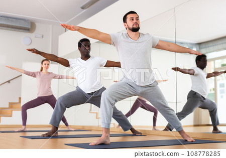 Bearded man standing in Virabhadrasana with group in yoga studio Bearded man standing in Virabhadrasana with group in yoga studio 108778285