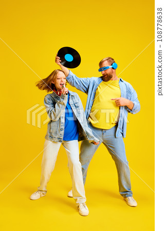 Father and daughter, parent and teenager in style denim outfit have fun with retro music plate and sing to microphone isolated yellow background. Father and daughter, parent and teenager in style denim outfit have fun with retro music plate and sing to microphone isolated yellow background. 108778638