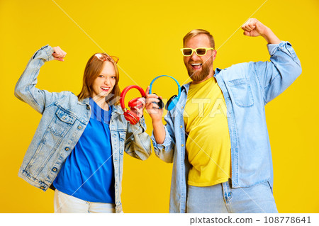 Funny father and daughter, parent and kid dressed style denim outfit and have fun holding headphones isolated yellow studio background. 108778641