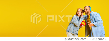Banner. Father and daughter, parent and kid dressed denim outfit and have fun listening music from one headphones over yellow background. Banner. Father and daughter, parent and kid dressed denim outfit and have fun listening music from one headphones over yellow background. 108778642