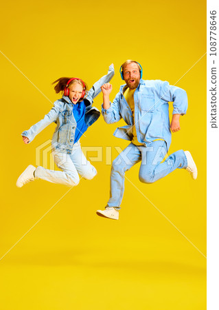 Father and daughter, parent and kid dressed style denim outfit and have fun listening music in headphones and jumping of joy isolated yellow background. 108778646