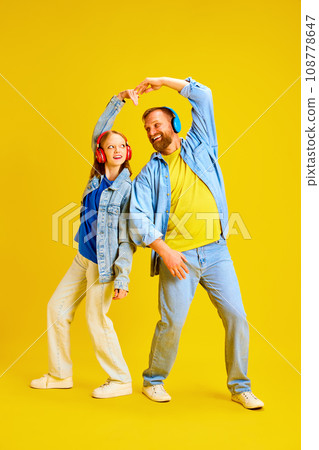 Father and daughter, parent and teen dressed style denim outfit and make hear from arms while listening music in headphones isolated yellow background. Father and daughter, parent and teen dressed style denim outfit and make hear from arms while listening music in headphones isolated yellow background. 108778647