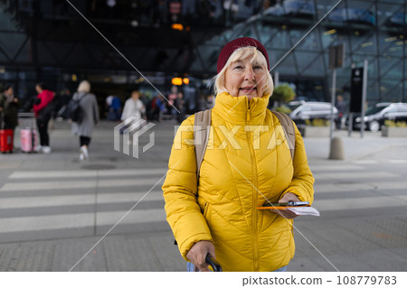 Happy attractive 60s senior female traveler walking with a suitcase and backpack at the modern transport stop outdoors near the modern airport terminal 108779783