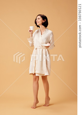 Young pretty lady in silk bathrobe drinking cup of hot coffee or tea before hard working day isolated beige studio background. Young pretty lady in silk bathrobe drinking cup of hot coffee or tea before hard working day isolated beige studio background. 108780121