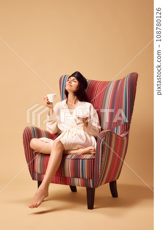 Young pretty lady sitting in armchair in silk bathrobe with cup of hot coffee or tea isolated beige studio background. 108780126