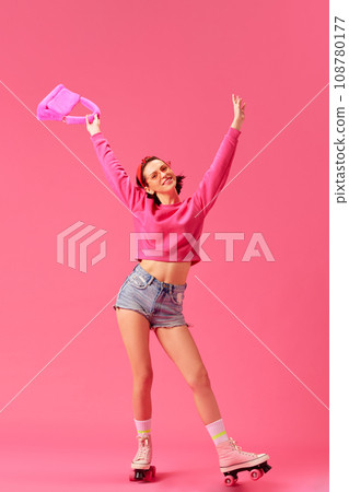 Excited pretty woman dressed shorts and vivid sweater stands on vintage roller skates and raising hands up of joy isolated pink studio background. 108780177